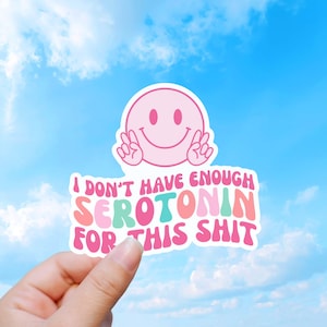 May include: A die-cut sticker featuring a light pink smiley face making a peace sign with both hands above the text I DON'T HAVE ENOUGH SEROTONIN FOR THIS SHIT written in a wavy, multi-colored font of pink, coral, and mint green against a white border.