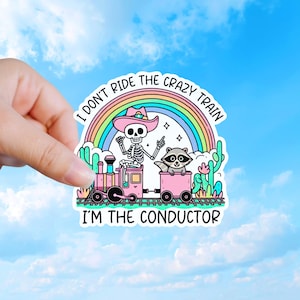 May include: A die-cut sticker featuring a skeleton wearing a pink cowboy hat sitting on a pink train engine, with a raccoon in a pink train car. A rainbow, cacti, and the text I DON'T RIDE THE CRAZY TRAIN I'M THE CONDUCTOR are included in the design.