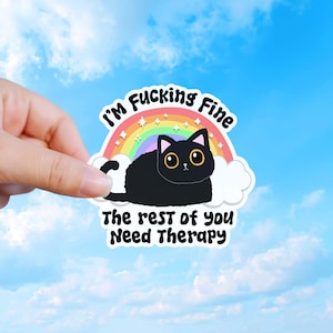 May include: A die-cut sticker featuring a black cat with large eyes sitting in front of a colorful rainbow and white clouds. The text reads I'M FUCKING FINE above the rainbow and THE REST OF YOU NEED THERAPY below the cat in black lettering.