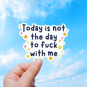 May include: A white die-cut sticker with a dark blue sans-serif text that reads Today is not the day to fuck with me. The sticker is decorated with small yellow and pink four-pointed star shapes scattered around the perimeter of the text.