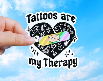 May include: A white sticker in the shape of a heart with the words "Tattoos are my Therapy" in black. The heart is filled with various black illustrations, including a skull, lightning bolt, and a bandage with rainbow stripes.