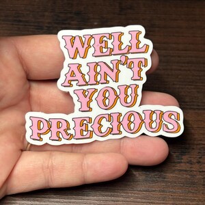 May include: A white sticker with the text "WELL AIN'T YOU PRECIOUS" in pink letters with orange outlines. The sticker is held in a hand against a dark brown wooden surface.