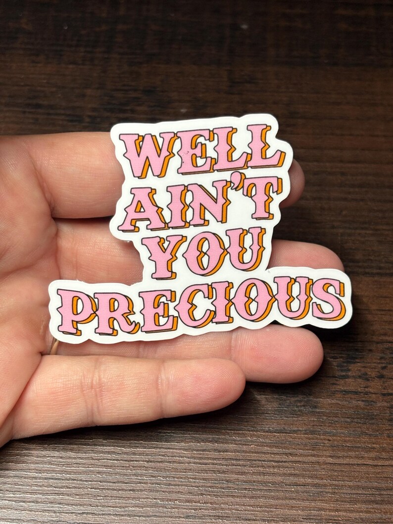 May include: A white sticker with the text "WELL AIN'T YOU PRECIOUS" in pink letters with orange outlines. The sticker is held in a hand against a dark brown wooden surface.