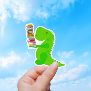 Könnte beinhalten: Ein gestanzter Aufkleber mit einem hellgrünen Cartoon-Dinosaurier, der einen hohen, vertikalen Stapel bunter Bücher hält. Der Dinosaurier hat einen runden Körper, einen kleinen Schwanz und dunkelgrüne Flecken auf dem Rücken sowie Bücher mit verschiedenfarbigen Buchrücken.