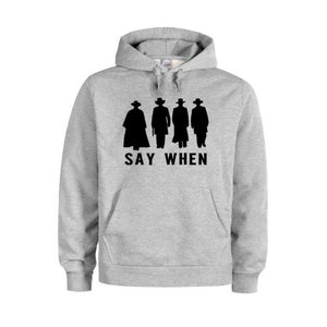 Puede incluir: Una sudadera con capucha gris con una silueta negra de cuatro figuras con sombreros y el texto "SAY WHEN" impreso en la parte delantera.