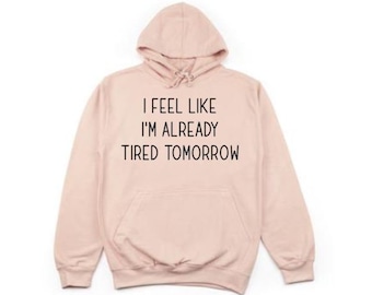 May include: A light pink hoodie with a drawstring hood and a front pocket. The text "I FEEL LIKE I'M ALREADY TIRED TOMORROW" is printed in black on the front. The hoodie is made of a soft material.