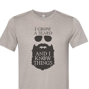 Puede incluir: Una camiseta gris jaspeada con un gráfico negro de una barba y gafas de sol. El texto en la camiseta dice "I grow a beard and I know things."
