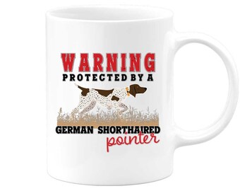 Könnte beinhalten: Weiße Keramik-Tasse mit Henkel. Auf der Tasse steht in Rot und Schwarz der Text "WARNING PROTECTED BY A GERMAN SHORTHAIRED POINTER". Eine Grafik eines Deutsch Kurzhaar Hundes ist ebenfalls auf der Tasse.
