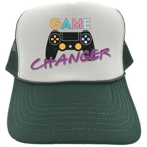 Könnte beinhalten: Grün-weiße Trucker-Cap mit dem Schriftzug "GAME CHANGER" und einer Videospiel-Controller-Grafik. Die Cap hat einen Mesh-Rücken und einen gebogenen Schirm. Der Text ist in einer farbenfrohen Retro-Schriftart.