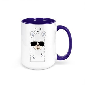 May include: White ceramic mug with a purple interior and handle. The mug features a cartoon llama wearing sunglasses, with the word "SUP" above the llama's head. A fun and quirky design for coffee or tea.