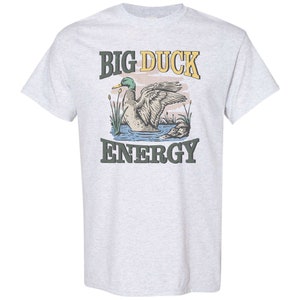 May include: A light grey t-shirt with a graphic of a duck in a pond and the text "BIG DUCK ENERGY".