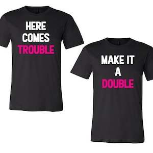 Op de afbeelding: Twee zwarte T-shirts met witte tekst. Het eerste shirt zegt "Here Comes Trouble" in wit met "Trouble" in roze. Het tweede shirt zegt "Make It A Double" in wit met "Double" in roze.