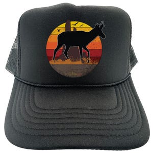 May include: Black trucker hat with a circular graphic featuring a black silhouette of a deer against a sunset backdrop of orange, red, and yellow stripes. The hat has a mesh back and a curved brim.
