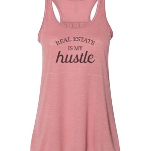 May include: A pink racerback tank top with the text "REAL ESTATE IS MY hustle" printed on the front.
