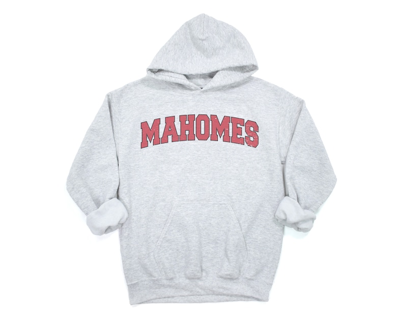 May include: A light gray hooded sweatshirt with the word "MAHOMES" in red, arched lettering across the chest. The sweatshirt has a front pocket and a drawstring hood. The sleeves are cuffed.
