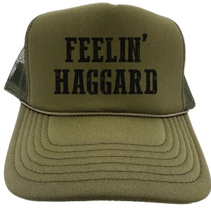 May include: A green trucker hat with a black mesh back. The front of the hat has the text "FEELIN' HAGGARD" in black.