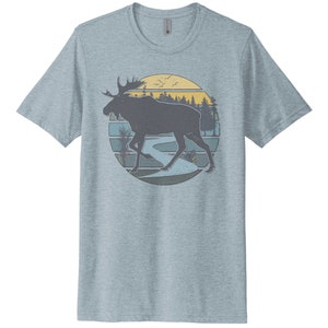 May include: A light blue t-shirt with a vintage-style graphic of a moose walking through a forest scene with a river and sunset in the background.