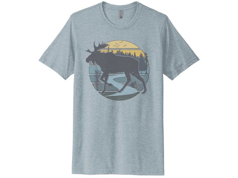 May include: A light blue t-shirt with a vintage-style graphic of a moose walking through a forest scene with a river and sunset in the background.