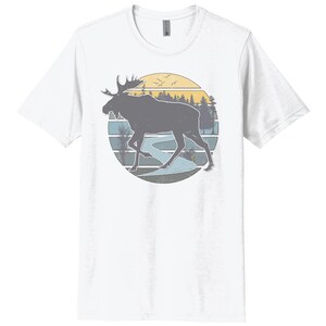 May include: A white t-shirt with a vintage-style graphic of a moose walking through a forest with a river in the background. The graphic is in shades of blue, yellow, and brown.