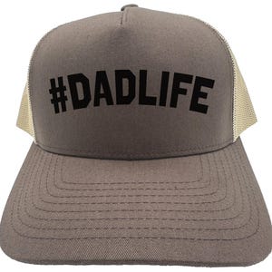 May include: A gray trucker hat with a mesh back and a curved brim. The front of the hat features the text "#DADLIFE" in bold, black letters. The hat is designed for casual wear.