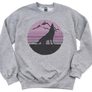 Puede incluir: Una sudadera gris con un gráfico desgastado de la silueta de un lobo aullando contra un atardecer rosa y púrpura con montañas. El diseño está dentro de un círculo. La sudadera tiene mangas largas y cuello redondo.