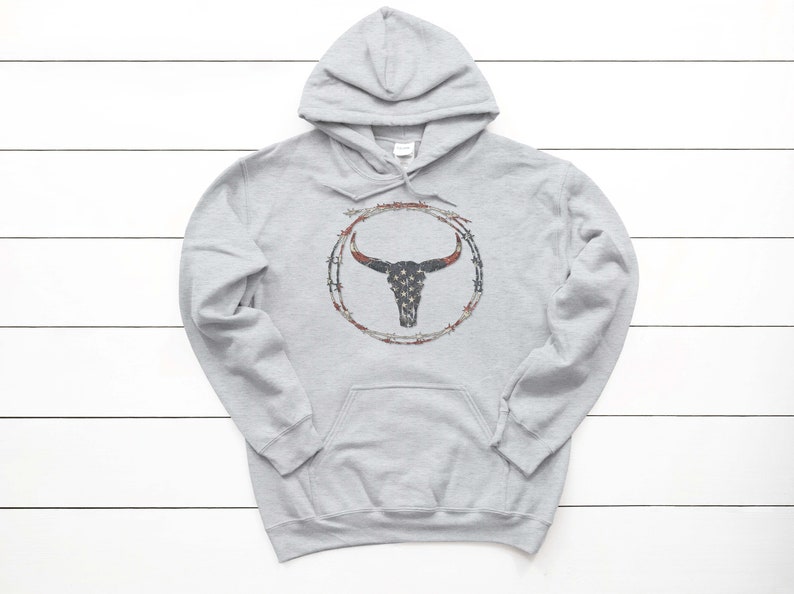 Puede incluir: Una sudadera con capucha gris con un dise&ntilde;o de cr&aacute;neo de toro. El cr&aacute;neo est&aacute; rodeado de alambre de p&uacute;as y tiene un patr&oacute;n de bandera estadounidense.
