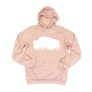 May include: A light pink hoodie with a white graphic design on the front. The hoodie has a drawstring hood and a kangaroo pocket. The graphic is a white shape with a grass-like design at the bottom.