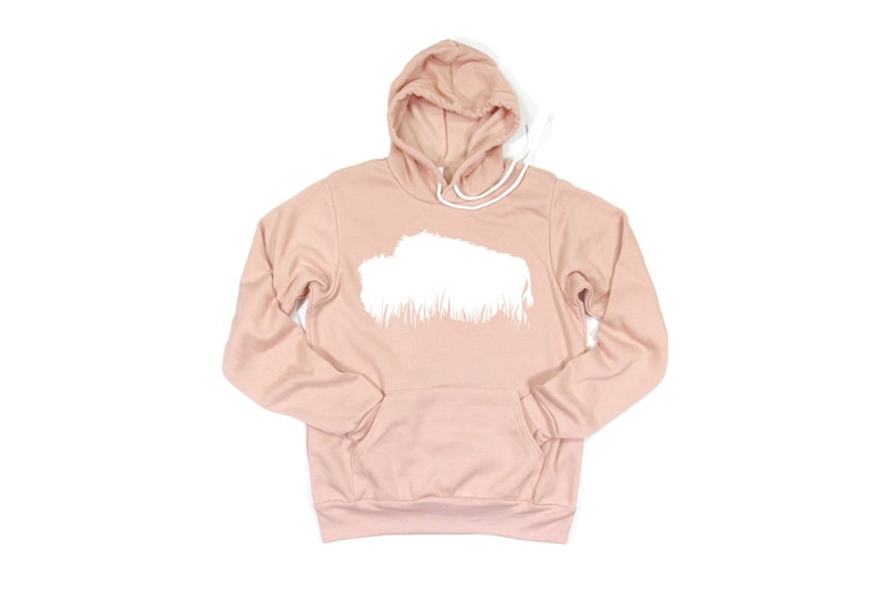 May include: A light pink hoodie with a white graphic design on the front. The hoodie has a drawstring hood and a kangaroo pocket. The graphic is a white shape with a grass-like design at the bottom.