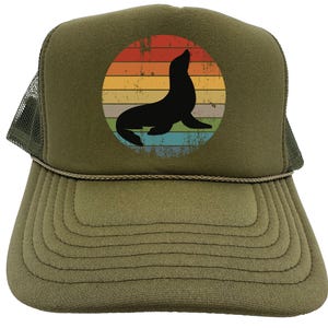 May include: A green trucker hat with a vintage-style sunset graphic featuring a silhouette of a seal. The hat has a mesh back and a brown adjustable strap.
