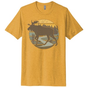 May include: A yellow t-shirt with a vintage-style illustration of a moose walking through a forest with a river in the background. The illustration is surrounded by a circle with a sunset behind the moose.