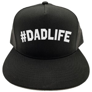May include: Black trucker hat with a flat brim and mesh back. The hat features the white text "#DADLIFE" across the front. The hat has a black crown and brim, with a gray mesh back panel. A black button is on top.