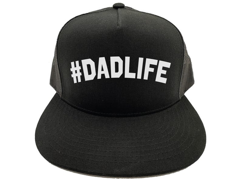 May include: Black trucker hat with a flat brim and mesh back. The hat features the white text "#DADLIFE" across the front. The hat has a black crown and brim, with a gray mesh back panel. A black button is on top.