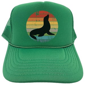 May include: A green trucker hat with a vintage-style graphic of a seal in a sunset. The seal is black and the sunset is in shades of orange, yellow, and red.