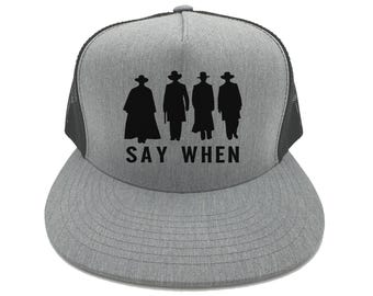 Puede incluir: Una gorra de camionero gris y negra con una silueta negra de cuatro figuras que llevan sombreros y abrigos. El texto "SAY WHEN" está impreso debajo de las figuras.