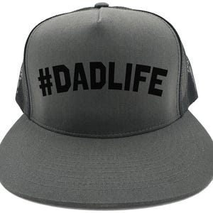 May include: A gray trucker hat with a black mesh back and a curved brim. The front of the hat features the text "#DADLIFE" in bold, black letters. The hat is a classic style, suitable for casual wear.