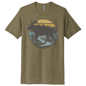May include: A green t-shirt with a vintage-style graphic of a moose walking through a forest with a river and sunset in the background.