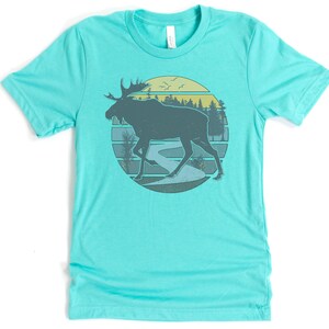 May include: A turquoise t-shirt with a retro-style graphic of a moose walking through a forest scene with a river and a sunset in the background.