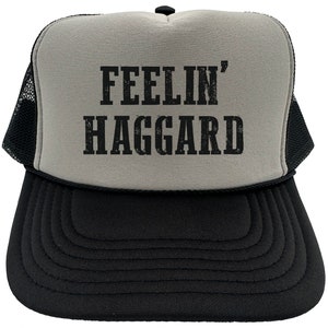 May include: A gray and black trucker hat with a black mesh back. The front of the hat has the text "FEELIN' HAGGARD" in black.
