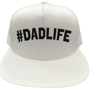 May include: White trucker hat with a curved brim and mesh back. The hat features the text "#DADLIFE" in bold, black letters across the front. A casual accessory for everyday wear.