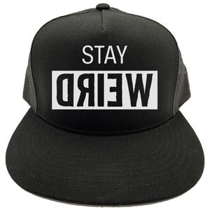 Può includere: Cappellino da camionista nero con visiera piatta e retro in rete. Il cappello presenta il testo bianco "STAY" sopra un rettangolo bianco con la parola "WEIRD" in grassetto maiuscolo. Un accessorio alla moda.