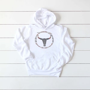 Puede incluir: Sudadera con capucha blanca con un gr&aacute;fico de un cr&aacute;neo de toro con un dise&ntilde;o de bandera estadounidense dentro de un c&iacute;rculo de alambre de p&uacute;as.