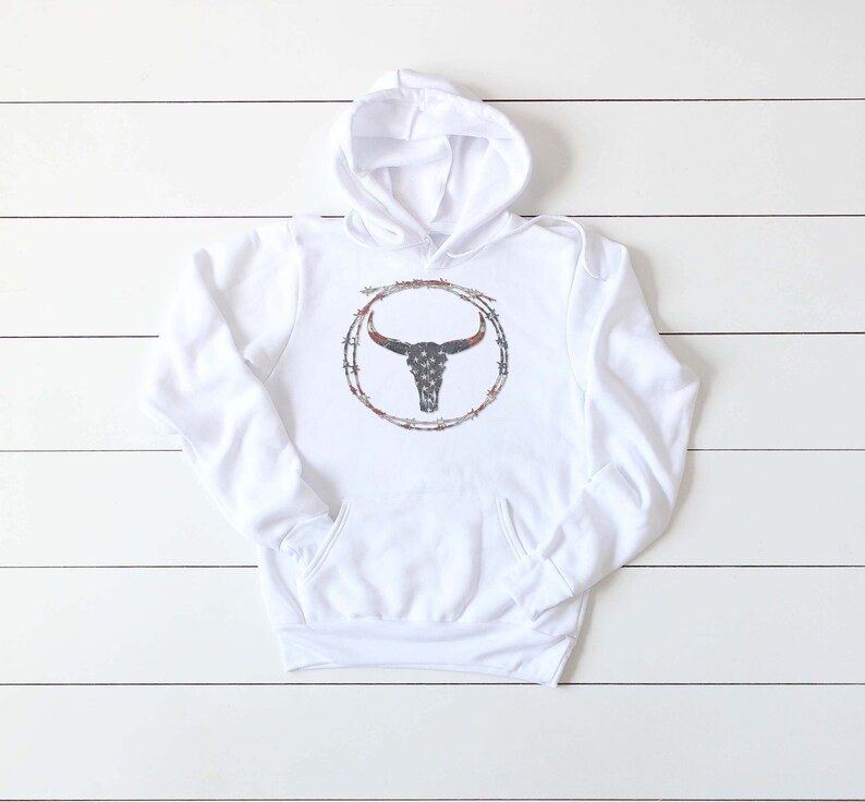 Puede incluir: Sudadera con capucha blanca con un gr&aacute;fico de un cr&aacute;neo de toro con un dise&ntilde;o de bandera estadounidense dentro de un c&iacute;rculo de alambre de p&uacute;as.
