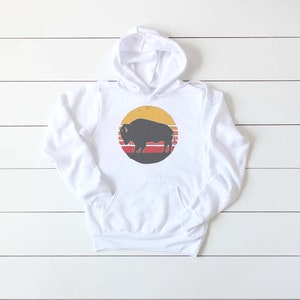 May include: White hooded sweatshirt with a retro-style graphic of a black silhouette of a bison in front of a yellow sun with red and orange stripes.
