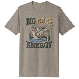 May include: A grey t-shirt with a vintage-style illustration of a duck swimming in a pond. The text "BIG DUCK ENERGY" is printed in yellow, green, and brown letters.