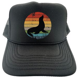 May include: Black trucker hat with a vintage-style graphic of a seal in front of a sunset. The graphic is in shades of orange, yellow, green, and blue.