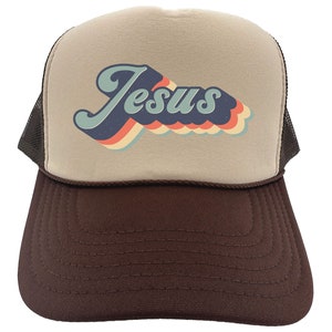Puede incluir: Una gorra de camionero marr&oacute;n y beige con un gr&aacute;fico retro de "Jes&uacute;s" en azul, rojo, naranja y amarillo.