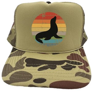May include: A brown and tan camouflage trucker hat with a vintage-style graphic of a seal in front of a sunset. The graphic is in a circle with a blue, green, and yellow color scheme.