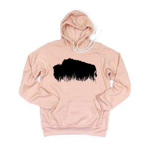 May include: A light pink hoodie with a black graphic of a bison. The hoodie has a drawstring hood and a front pocket. The bison graphic is a silhouette with a textured, grassy base.