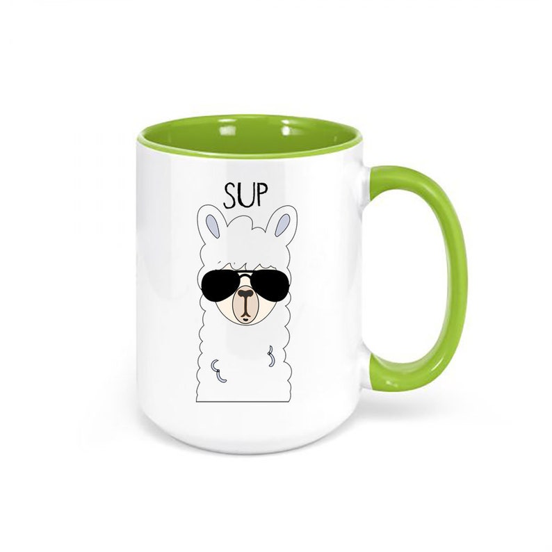 May include: White ceramic mug with a green handle and interior. The mug features a cartoon llama wearing sunglasses, with the word "SUP" above the llama's head. A fun and quirky design for a coffee or tea mug.