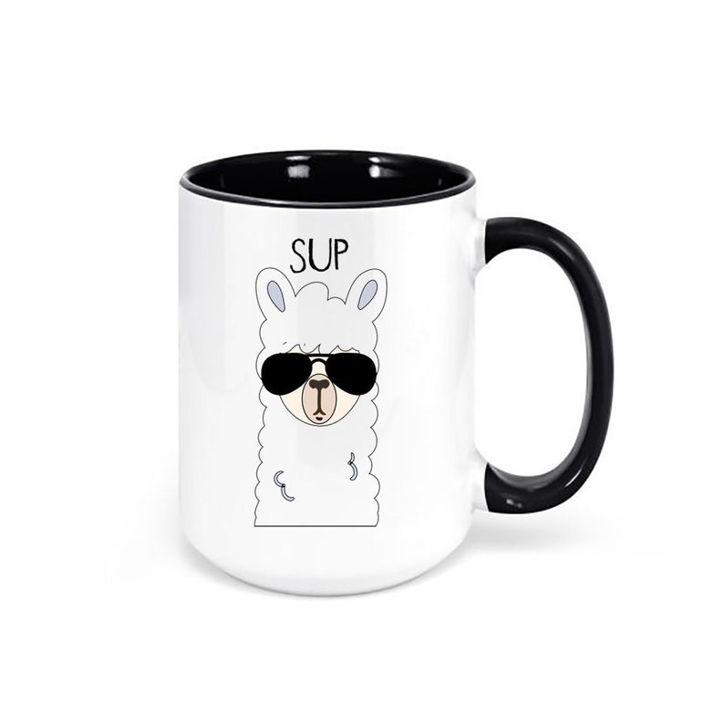 May include: White ceramic mug with a black interior and handle. The mug features a cartoon llama wearing sunglasses, with the word "SUP" above it. A fun and quirky design for coffee or tea.